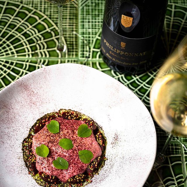 Plat gastronomique avec une mousse rose et des feuilles vertes sur une assiette blanche, avec une bouteille de vin et des verres en arrière-plan