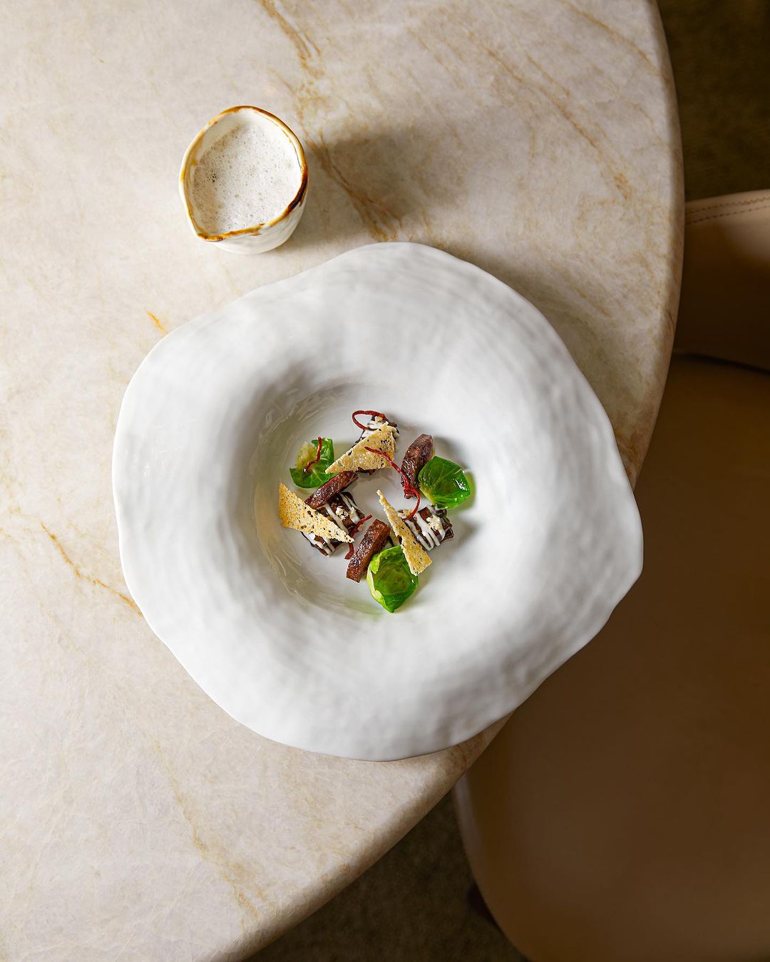 Plat gastronomique dans une assiette blanche texturée avec des légumes et des garnitures artistiques, accompagné d'un petit bol de sauce à côté.