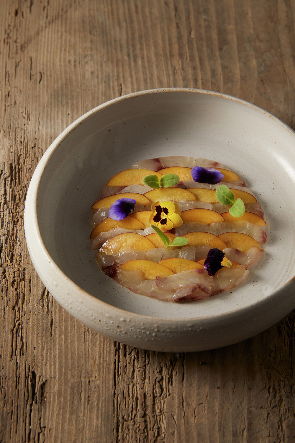 Carpaccio de fruits et de poisson cru, décoré de fleurs comestibles, servi dans un bol en céramique.