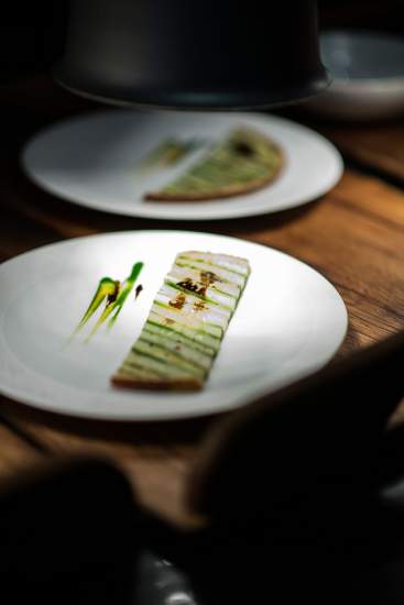 Plat gastronomique joliment présenté, éclairé sous une lampe, sur une assiette blanche avec des décorations vertes et jaunes.