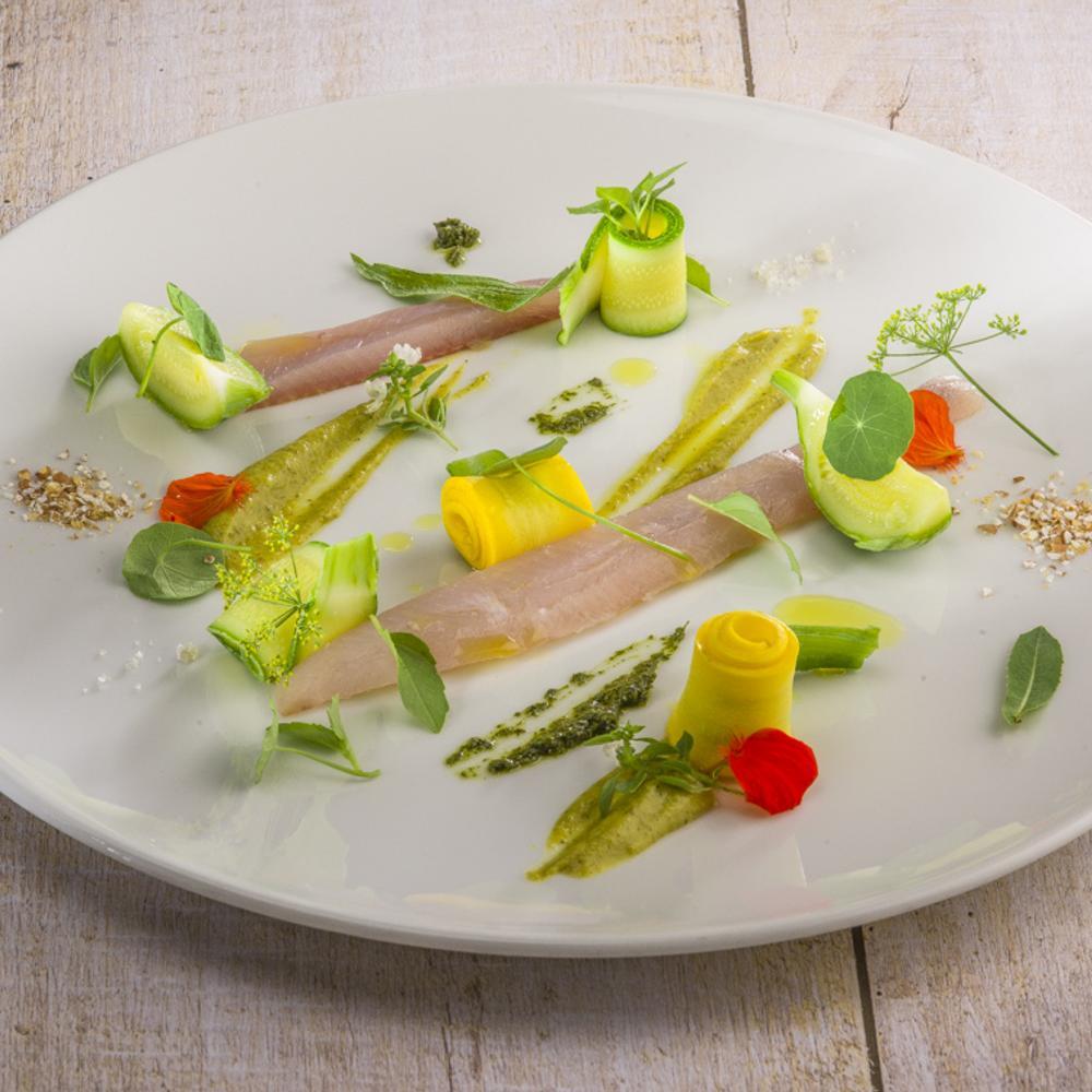 Assiette gastronomique avec des tranches de poisson cru, des rouleaux de légumes et des touches de sauces vertes et jaunes.
