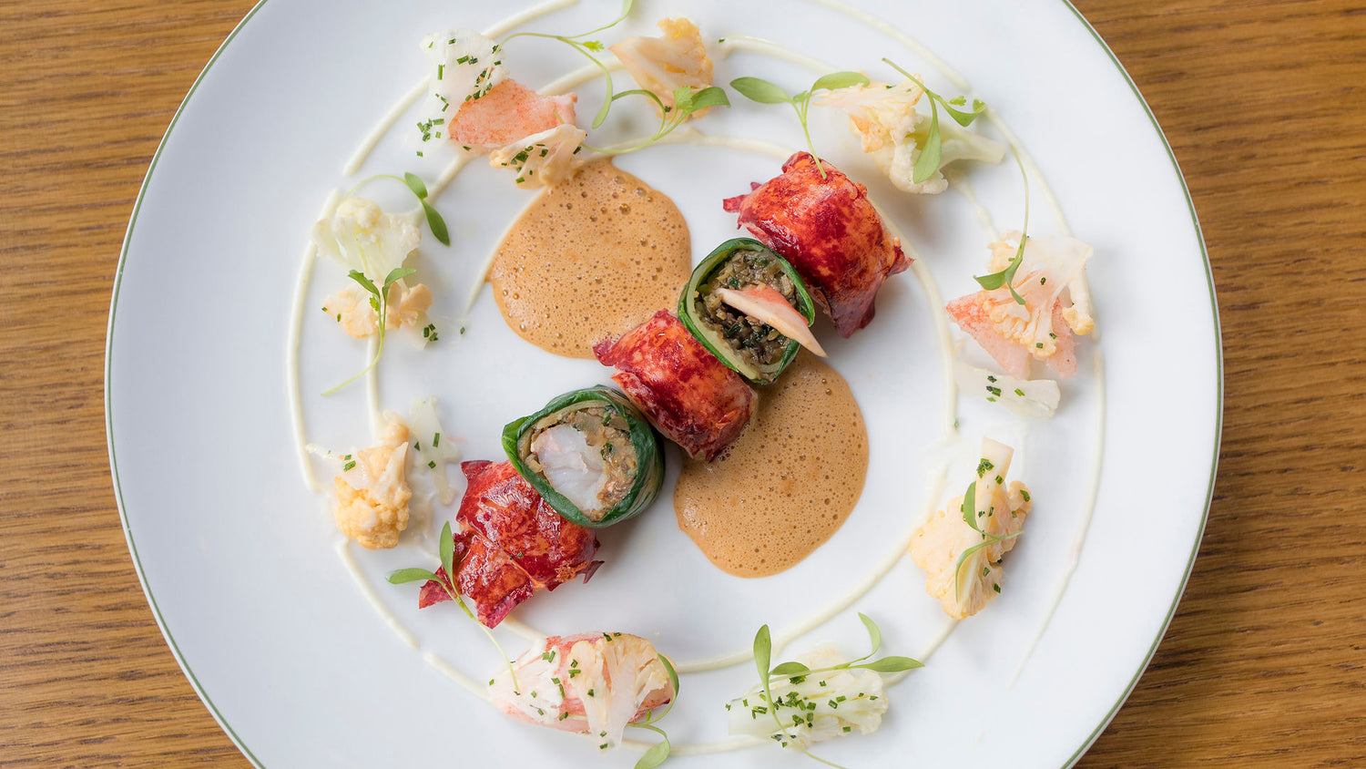 Rouleaux de homard enrobés de feuilles vertes, accompagnés de petites fleurs comestibles et d'une sauce légère, présentés sur une assiette élégante.