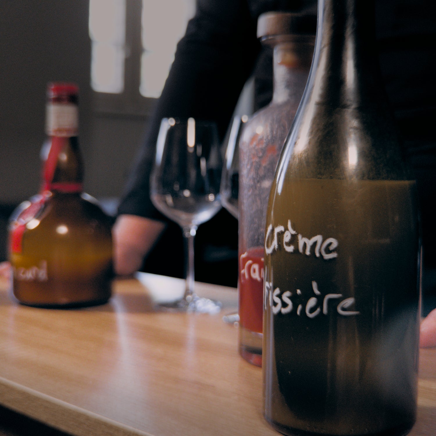 Gros plan sur des bouteilles de liqueur disposées sur un comptoir en bois, avec un verre à vin vide au centre. Une des bouteilles est étiquetée "Crème pâtissière". Une personne en arrière-plan semble préparer quelque chose. La lumière douce crée une ambiance intime et chaleureuse.