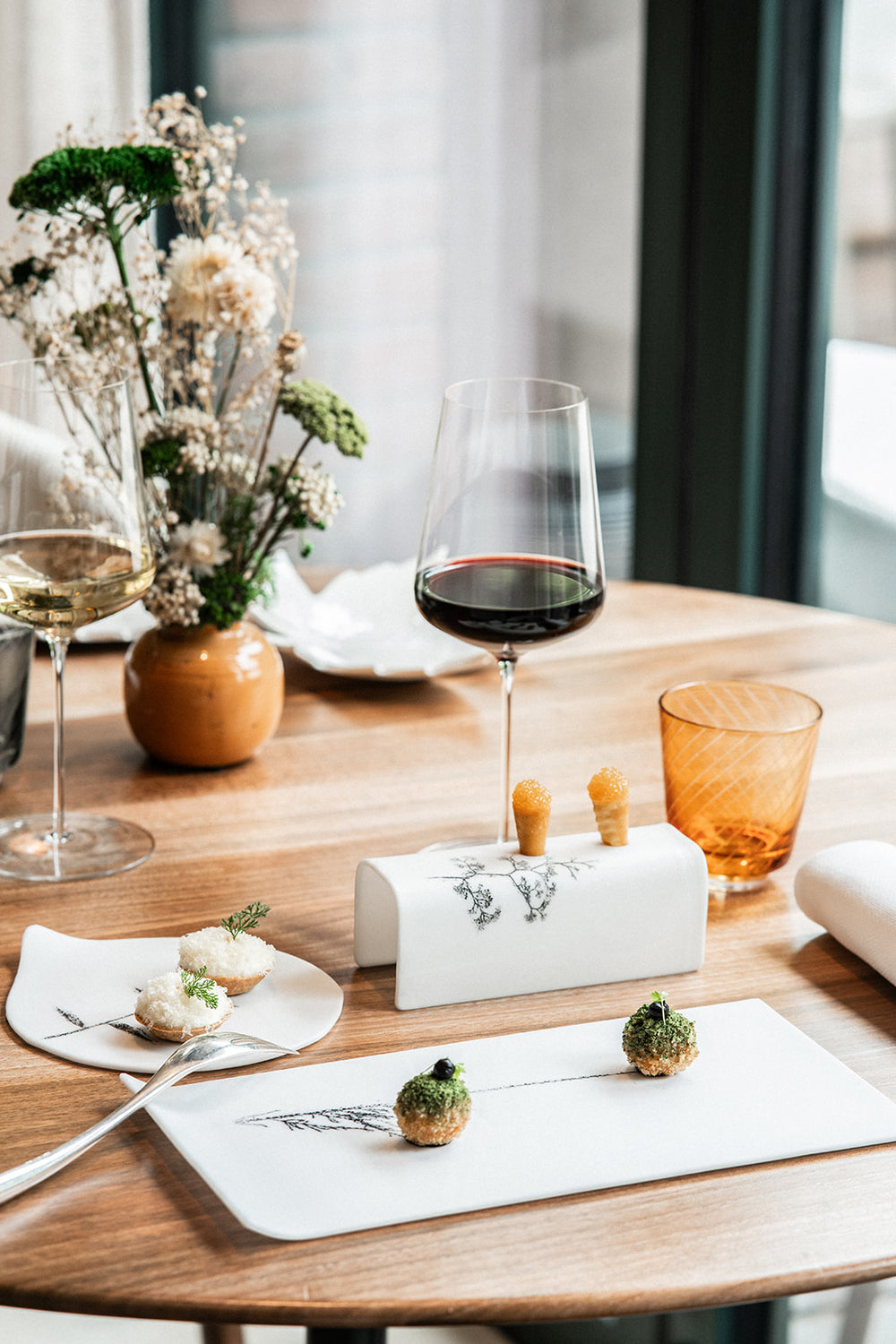 Table élégamment dressée avec des amuse-bouches raffinés présentés sur des assiettes artistiques, accompagnés de verres de vin rouge et blanc, dans une atmosphère lumineuse et fleurie.