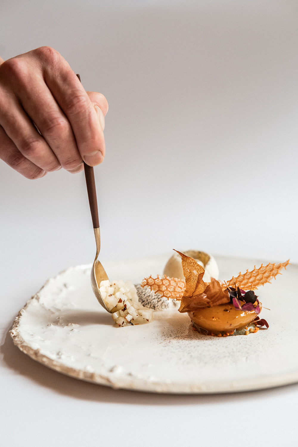 Main tenant une cuillère en train de déguster un plat gastronomique artistiquement dressé avec des textures croustillantes, des morceaux de légumes finement coupés et des éléments visuellement raffinés sur une assiette en céramique texturée blanche.