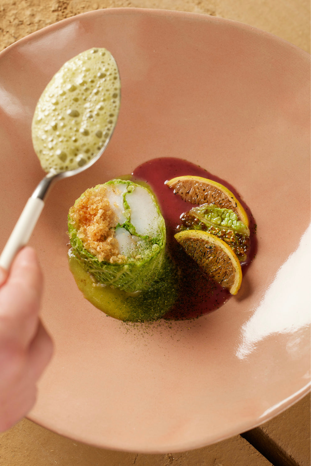 Un plat raffiné servi dans une assiette beige, avec un roulé de chou farci accompagné de quartiers d'agrumes et d'une émulsion crémeuse, versée délicatement à la cuillère.