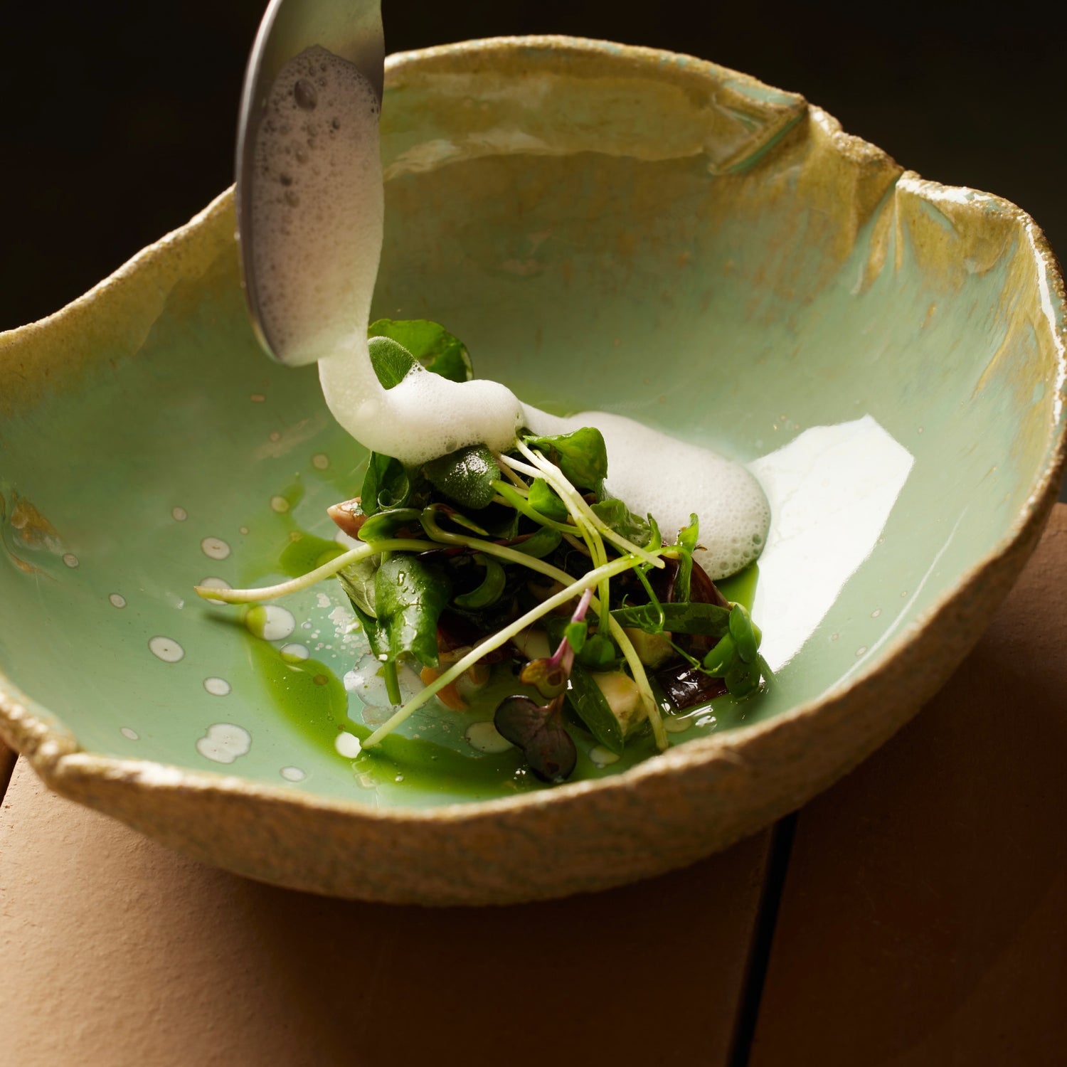 Un plat gastronomique délicatement préparé, garni de jeunes pousses et arrosé d'une émulsion crémeuse versée avec une cuillère dans un bol en céramique verte.