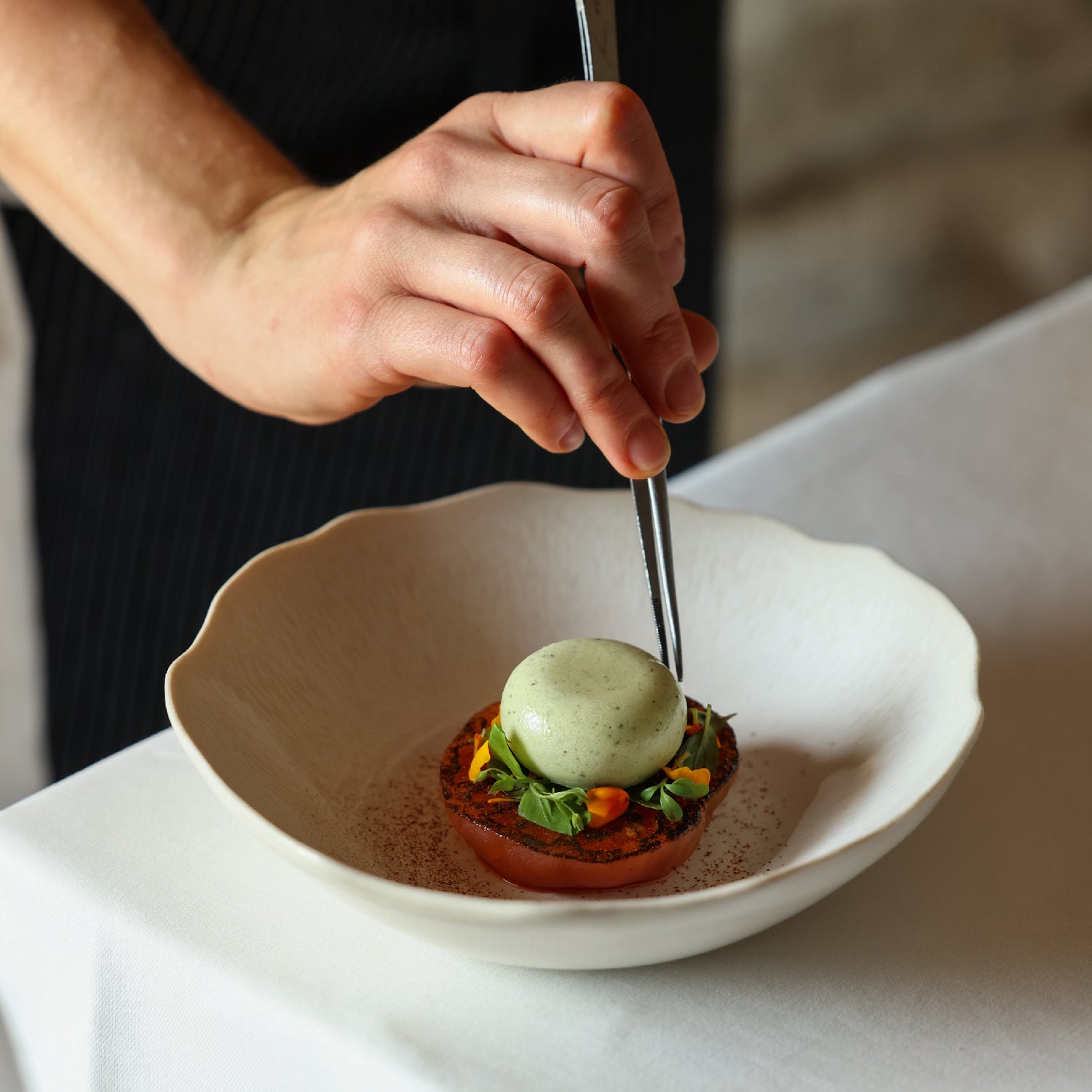 Plat raffiné composé d’une tomate grillée garnie de jeunes pousses et fleurs, surmontée d’une boule de crème verte, dressé avec précision à la pince.