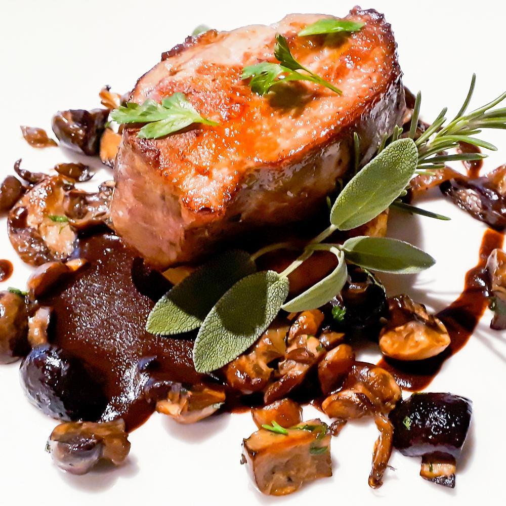 Pièce de viande grillée, servie avec une sauce riche et des champignons sautés, décorée de feuilles de sauge et de romarin, dans une présentation rustique et appétissante.