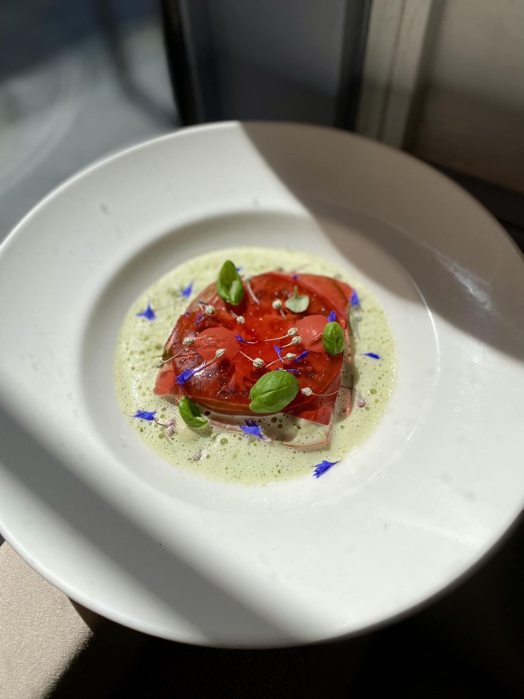 Carpaccio de tomates avec mousse verte et décor floral, servi dans une assiette blanche épurée baignée de lumière naturelle.