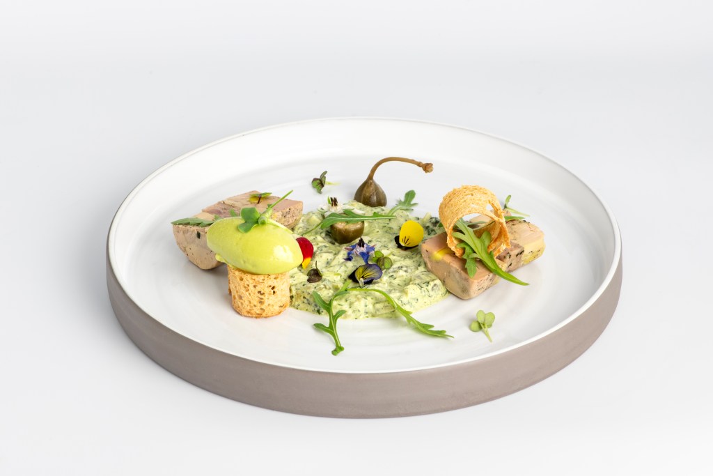 Assiette gastronomique comprenant diverses préparations élégamment disposées avec une mousse verte, des caprons, des fleurs comestibles, et des garnitures colorées.