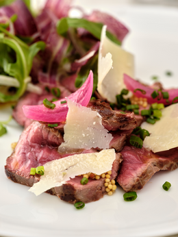 Plat gastronomique composé de tranches de viande rouge saignante, garnies de copeaux de parmesan, de pickles d'oignon rouge, de grains de moutarde, et d'herbes fraîches, accompagné d'une salade verte.