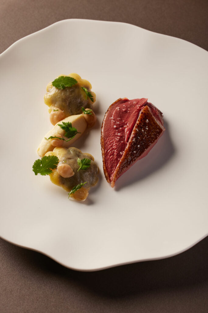 Un plat gastronomique avec un morceau de viande rosée soigneusement présenté sur une assiette blanche. À côté, des raviolis garnis, le tout décoré avec des herbes fraîches. La présentation est minimaliste mais raffinée, mettant en valeur chaque composant du plat.