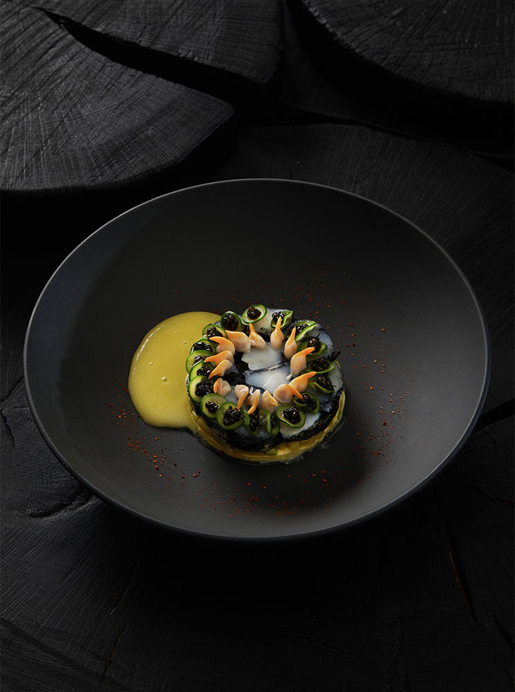 Un plat magnifiquement présenté avec une sauce jaune éclatante accompagnant des ingrédients finement disposés dans une assiette noire élégante, sur un fond de textures sombres.