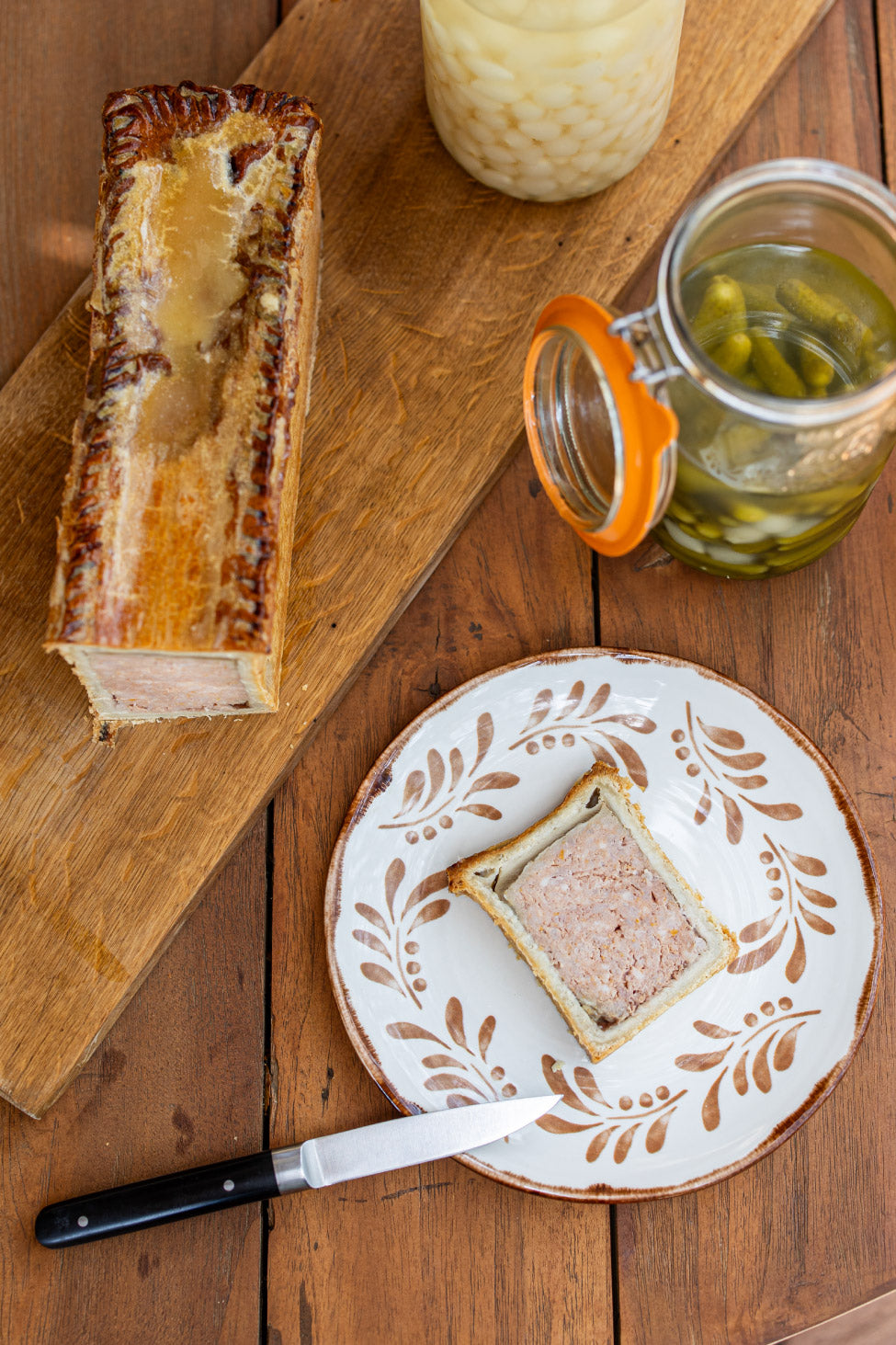 Vue de dessus d'un pâté en croûte sur une planche en bois, accompagné de cornichons dans un bocal et d'une assiette avec une tranche de pâté en croûte, posée sur une table en bois rustique.