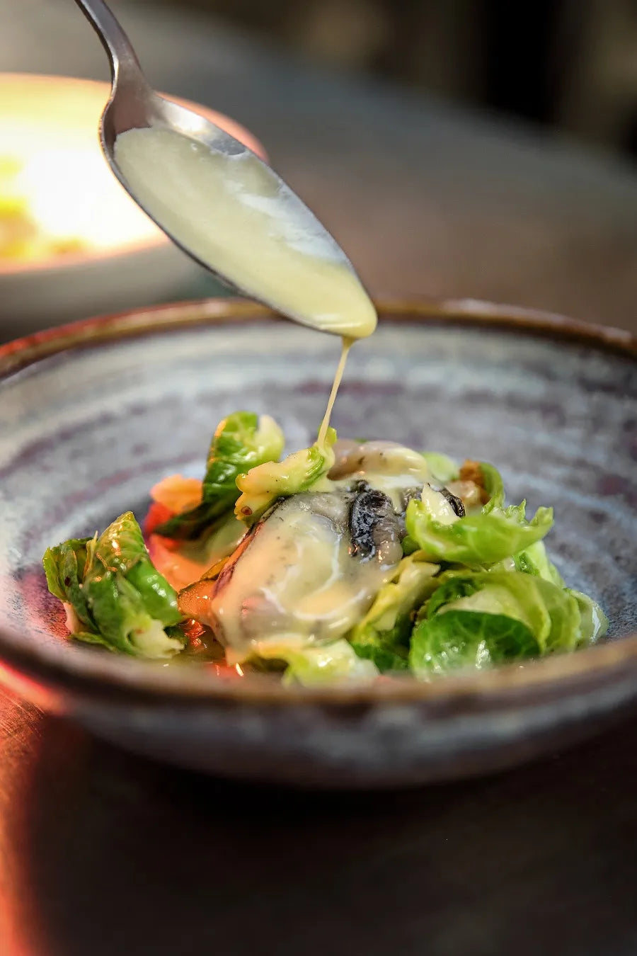 Sauce crémeuse versée sur un plat d'huîtres et légumes verts dans une assiette en céramique texturée