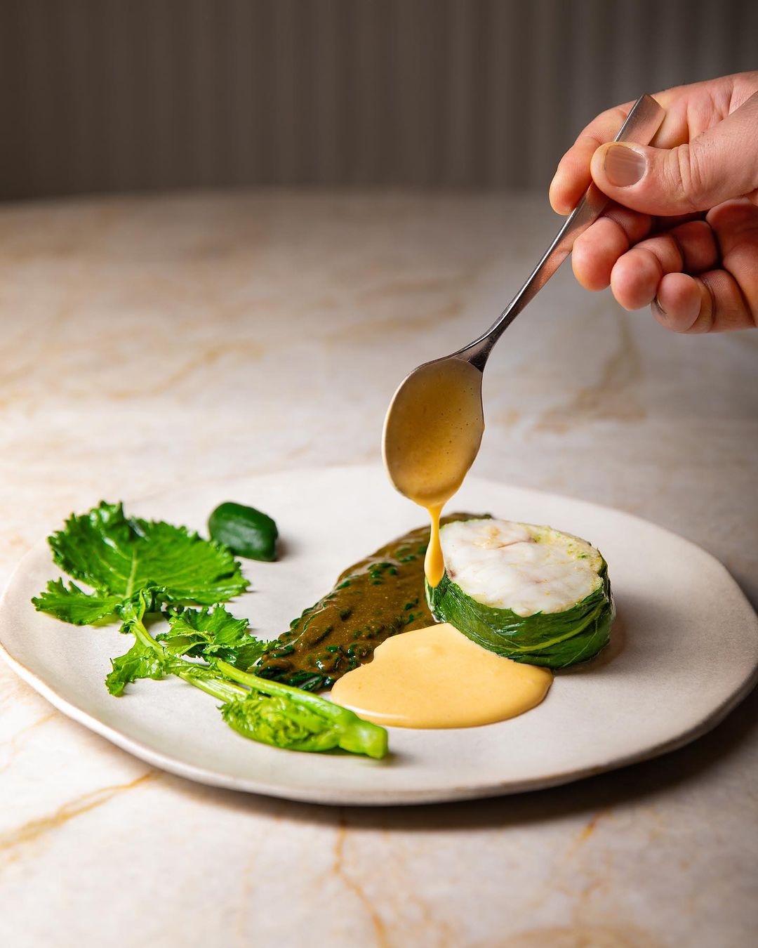 Main versant une sauce sur un plat élégant avec des légumes verts et du poisson enveloppé dans des feuilles.