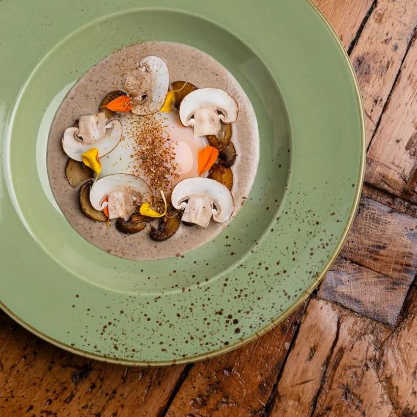 Plat gastronomique avec un œuf poché entouré de tranches de champignons et de garnitures colorées, présenté dans une assiette verte.