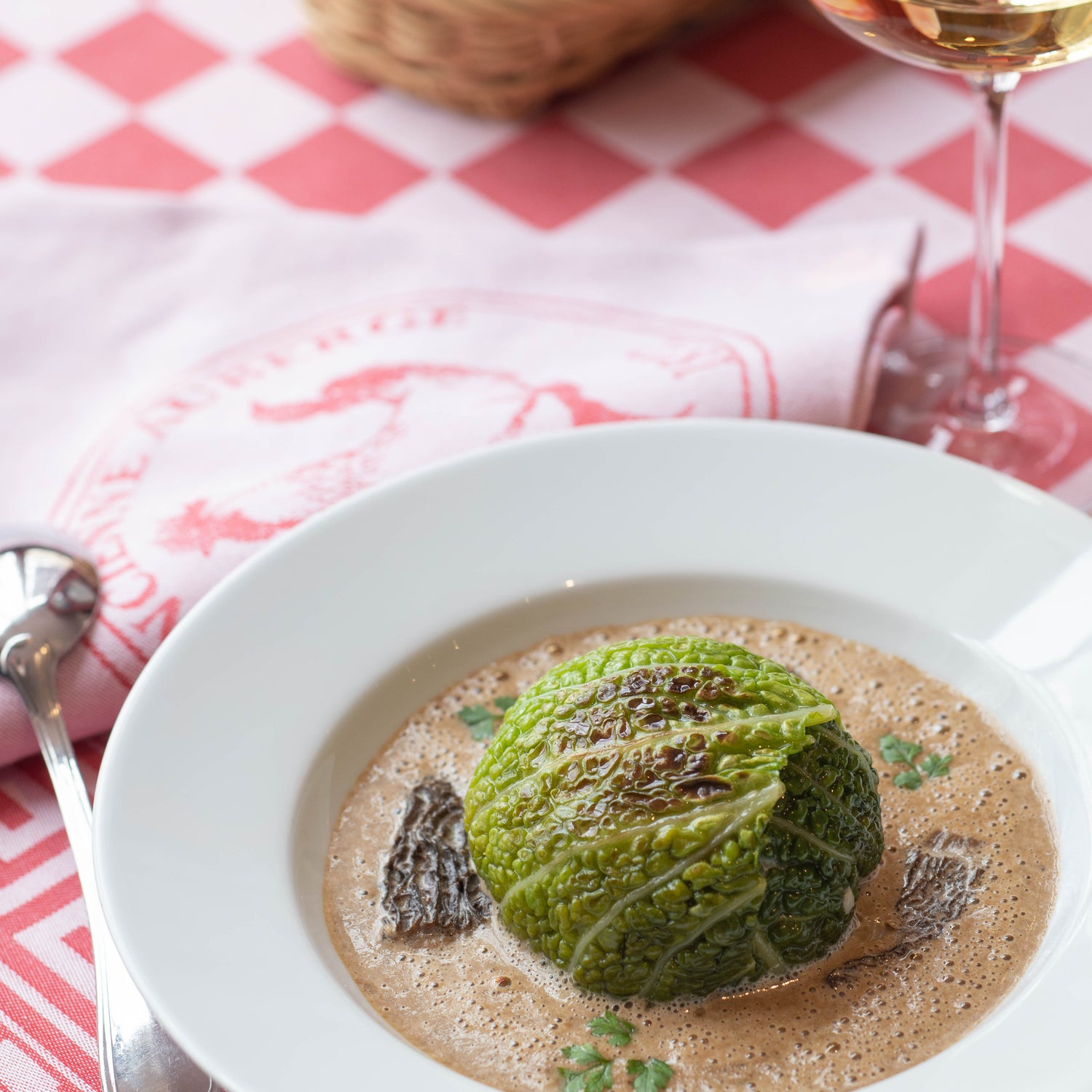 Chou farci servi dans une assiette blanche, accompagné d'une sauce crémeuse et de morilles, posé sur une nappe à carreaux rouges et blancs avec un verre de vin blanc à côté.