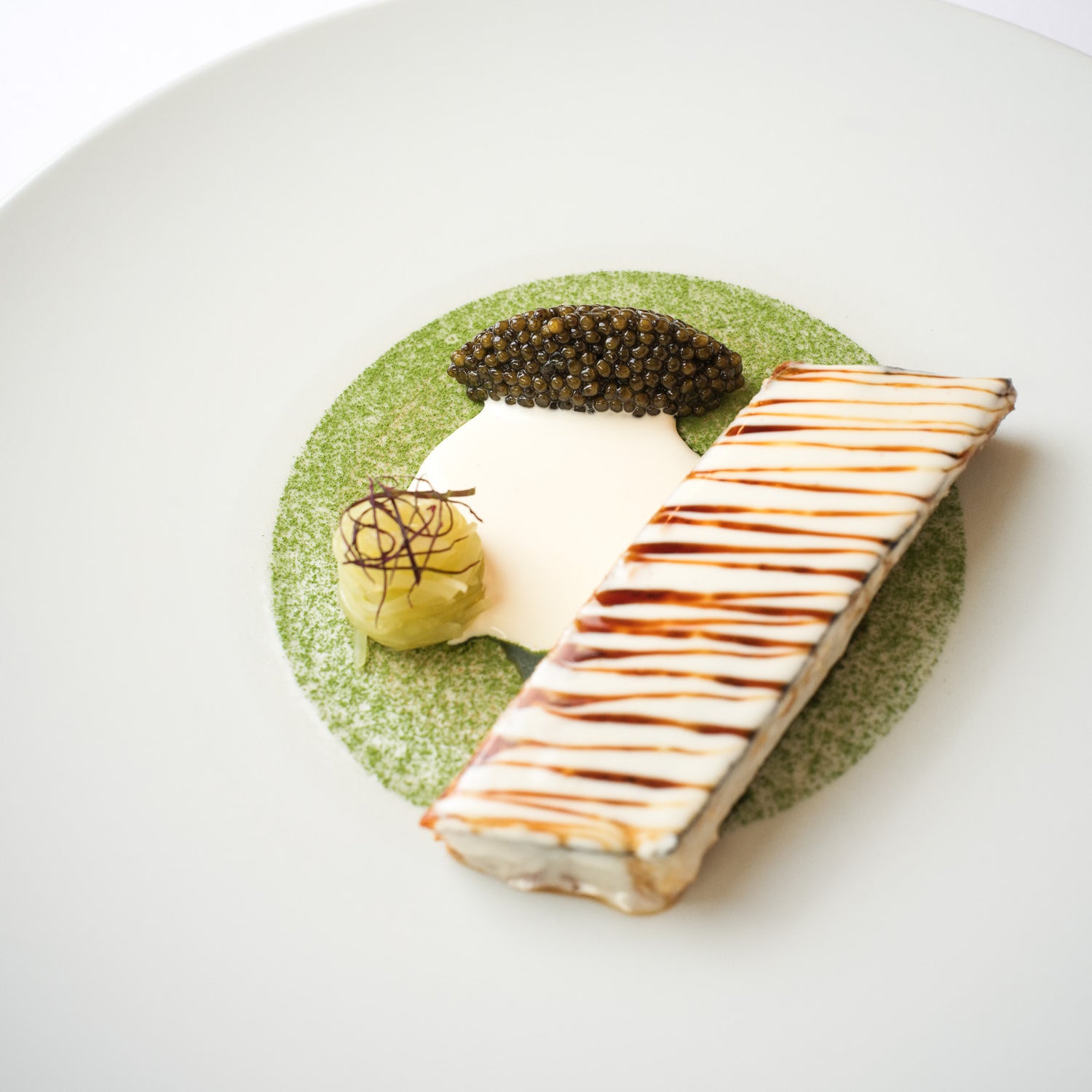 Élégant plat gastronomique avec une barre de poisson nappée de sauce, servie avec du caviar, une crème légère et une garniture délicate sur une assiette décorée de poudre verte.