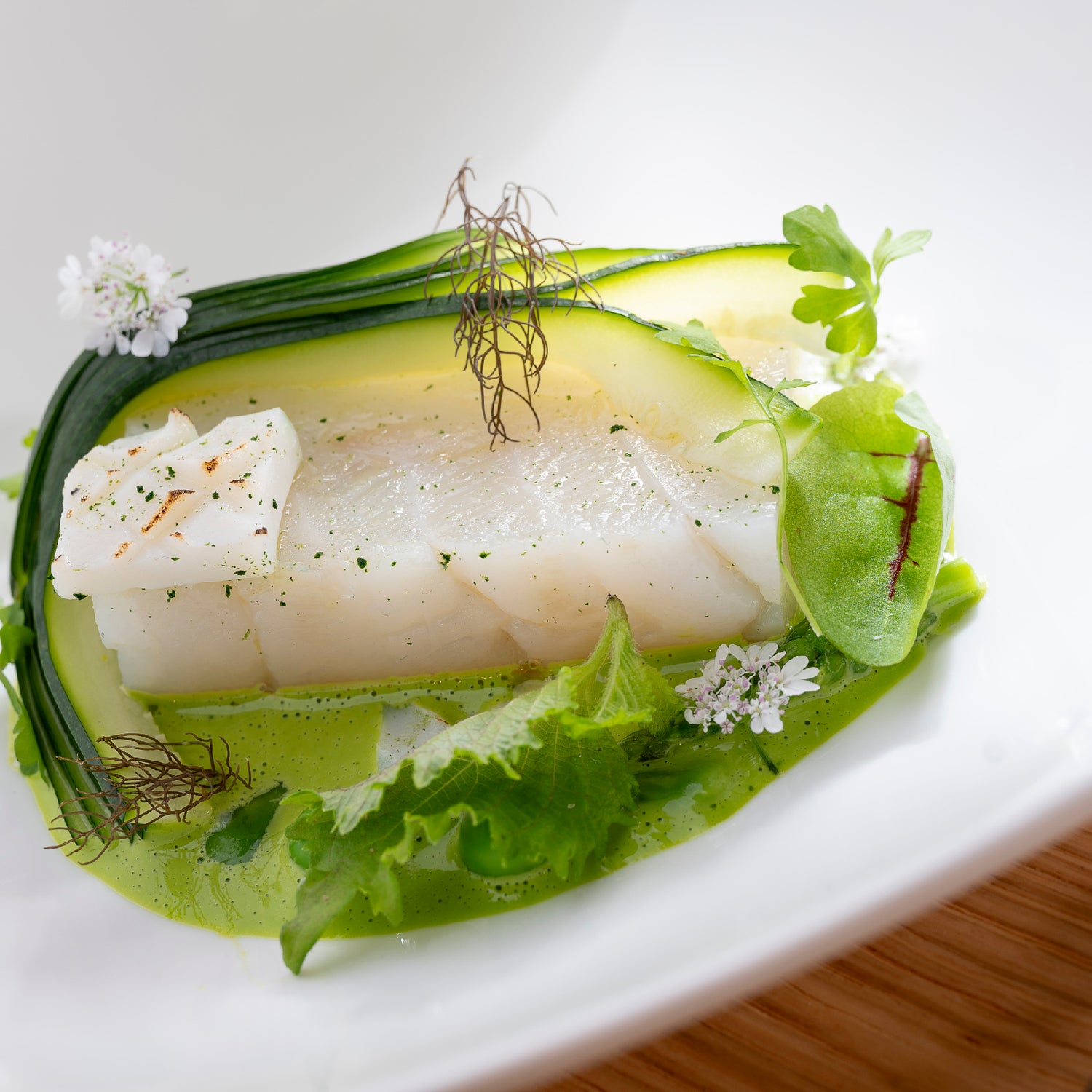 Filet de poisson blanc délicatement cuit, accompagné de courgettes finement tranchées, de verdure fraîche et de fleurs comestibles, servi dans une assiette blanche sur fond de bois clair.