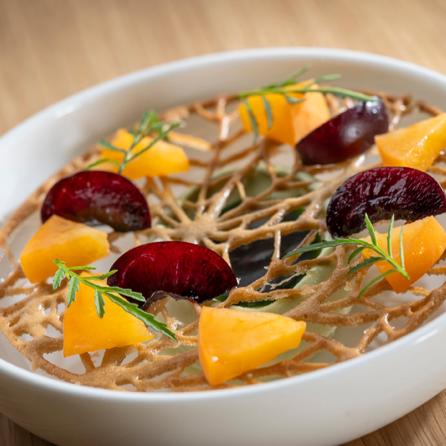 Dessert raffiné présenté dans une assiette blanche, composé d’un disque croustillant ajouré, garni de morceaux de fruits jaunes et rouges, et décoré de petites herbes fraîches, sur fond de bois clair.