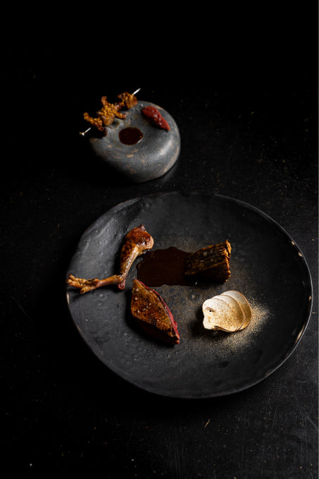 Assiette gastronomique élégante composée d’un pigeon rôti, accompagné d’une morille, d’une purée en spirale et d’une sauce brune, avec en arrière-plan une pierre de présentation garnie de bouchées croustillantes et d’un condiment.