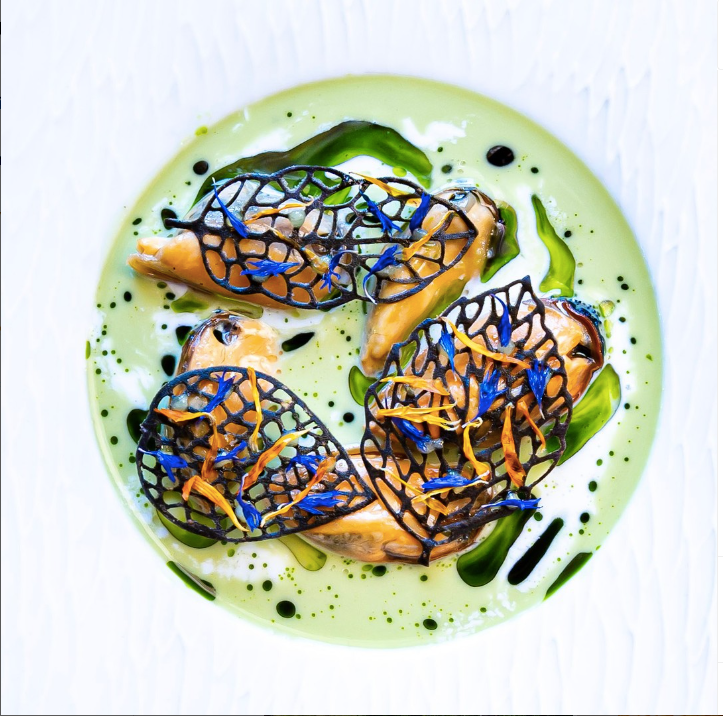 Une assiette gastronomique élégante présentant des moules accompagnées d'une décoration artistique de dentelle comestible noire, le tout disposé sur une sauce verte onctueuse avec des touches de couleur contrastantes.