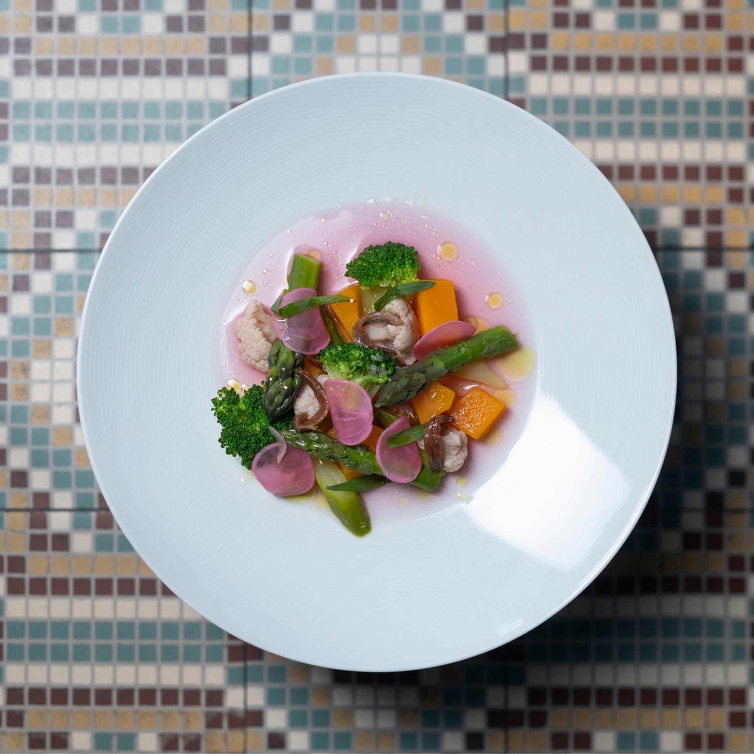 Plat gastronomique coloré présenté dans une assiette blanche, avec des légumes variés tels que des asperges, brocolis, radis et morceaux de potiron, sur un fond de sauce rose. Le tout est posé sur un sol en mosaïque aux motifs géométriques.