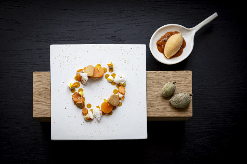 Dessert raffiné composé de petites touches de crème et de fruits disposées en cercle sur une assiette blanche, accompagné d'une quenelle de glace et d'une sauce, le tout présenté de manière élégante sur une table en bois.