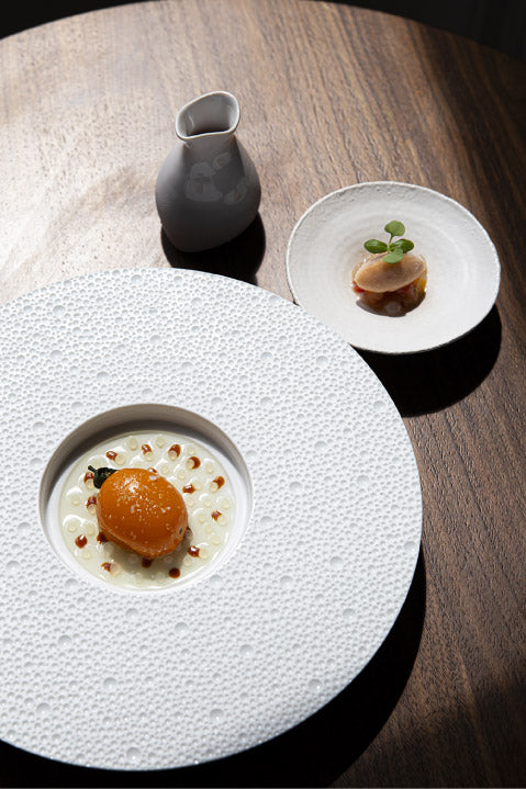 Présentation gastronomique d'un plat sur une assiette texturée blanche, accompagné d'une petite assiette et d'une carafe sur une table en bois. Le plat principal se compose d'une préparation raffinée avec une garniture délicate, tandis que la petite assiette présente une autre création culinaire élégante.
