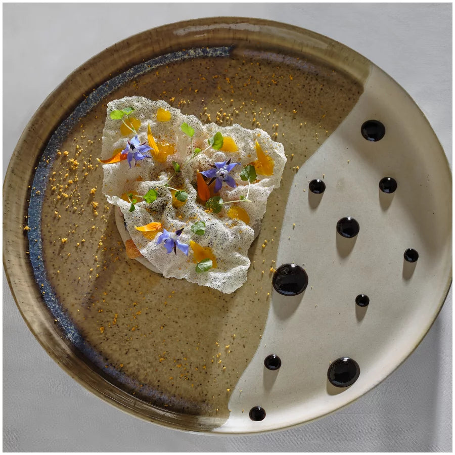 Assiette artistique avec dentelle comestible, fleurs colorées et gouttes de sauce noire.