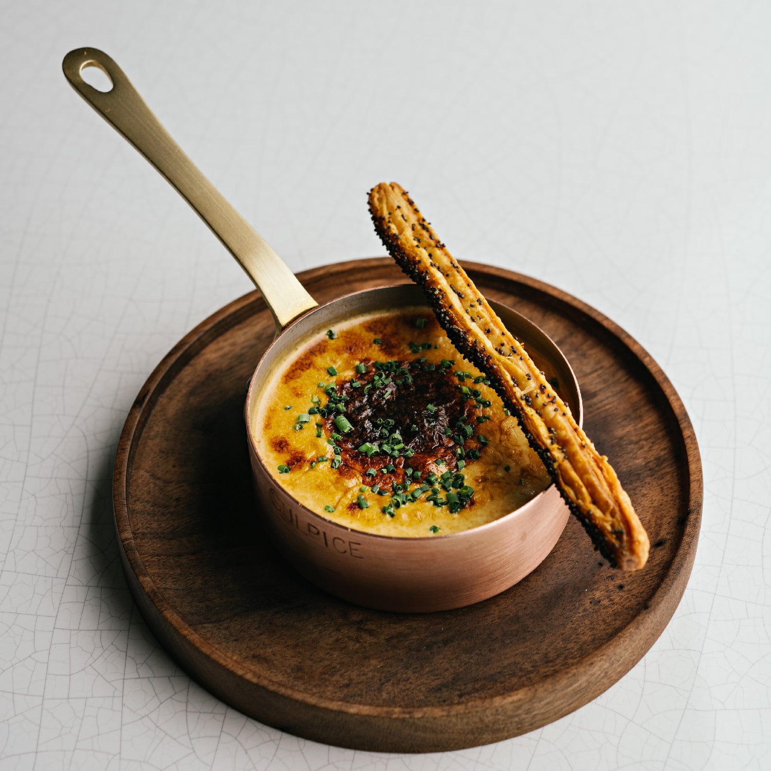 Un plat gastronomique servi dans une casserole en cuivre, garni de fines herbes et accompagné d'un bâtonnet feuilleté doré, présenté sur une assiette en bois.