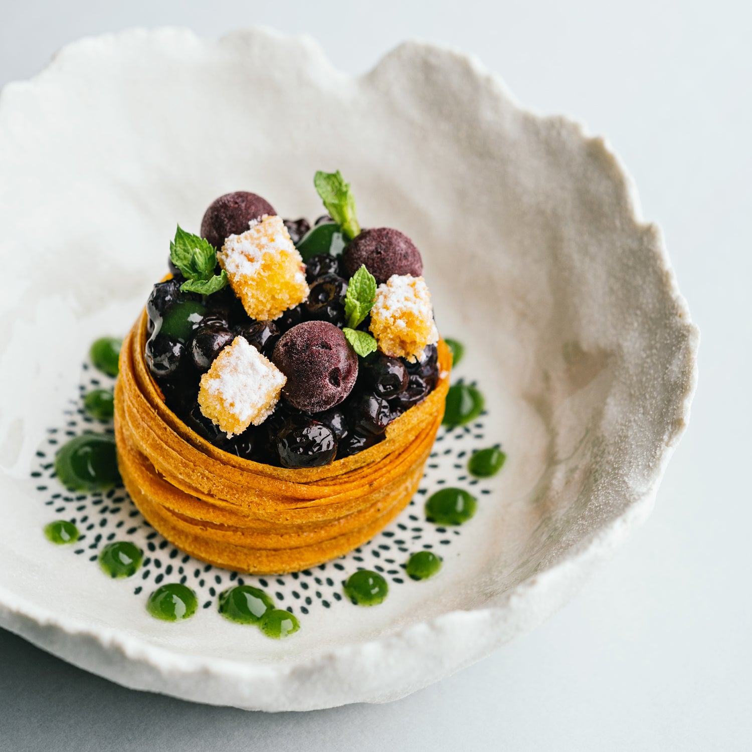 Dessert raffiné présenté dans une assiette en céramique, composé d'une base croustillante garnie de petites baies, de cubes de garniture dorée et saupoudrée de sucre, décorée de feuilles de menthe et entourée de points de coulis vert.