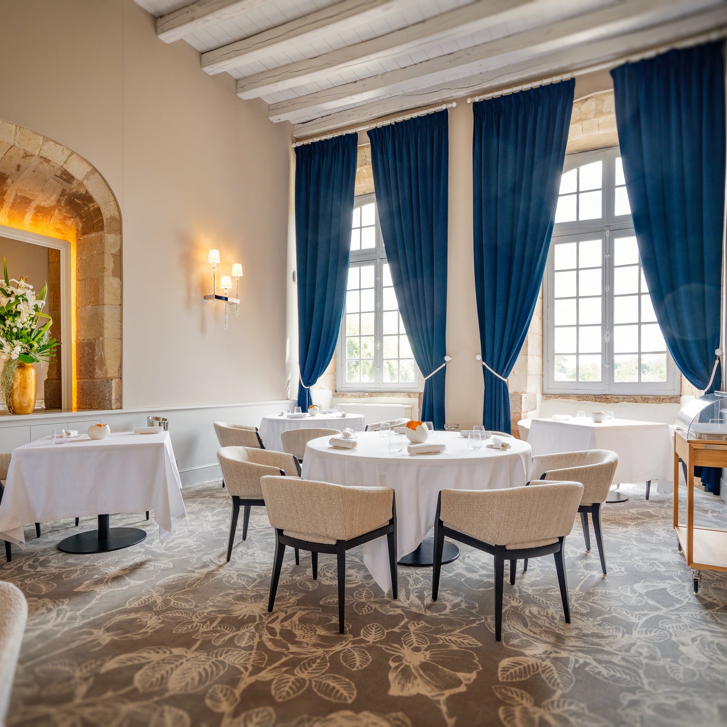 Salle de restaurant lumineuse et élégante avec des tables rondes et carrées dressées de nappes blanches, des chaises rembourrées confortables, des grandes fenêtres encadrées de rideaux bleus et une décoration florale raffinée.