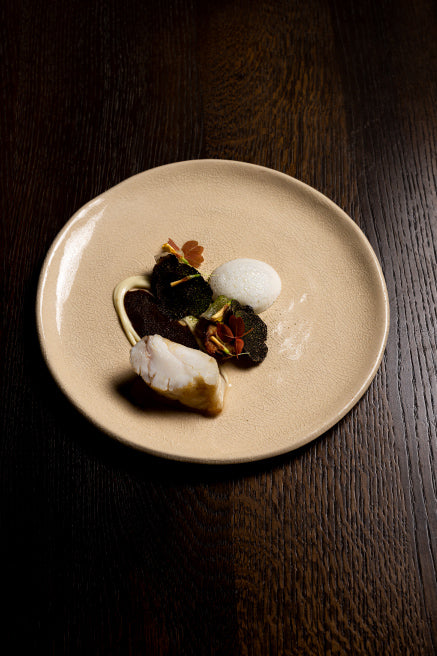 Assiette gastronomique raffinée composée d’un morceau de poisson blanc nappé de sauce, accompagnée de légumes noirs croustillants, de jeunes pousses et d’une écume légère, le tout dressé avec élégance sur une assiette beige.