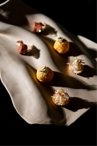 Assortiment de bouchées gastronomiques élégamment disposées sur un plateau en céramique ondulé, comprenant des choux garnis et décorés de fleurs comestibles et de fines herbes.