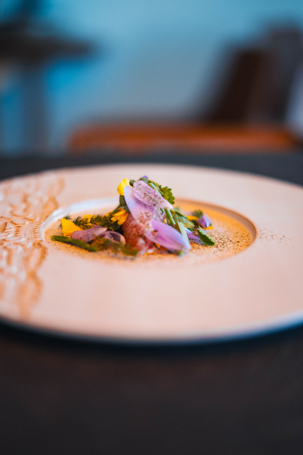 Plat gastronomique coloré dressé avec élégance, composé de légumes, herbes fraîches et fleurs comestibles, disposés au centre d’une assiette blanche ornée d’un motif texturé.