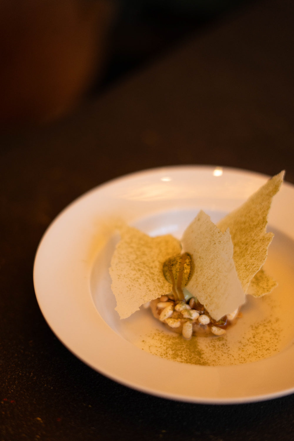 Dessert gastronomique raffiné présenté dans une assiette blanche, garni de tuiles croustillantes, d’herbes et de grains soufflés, avec une touche de poudre verte décorative.