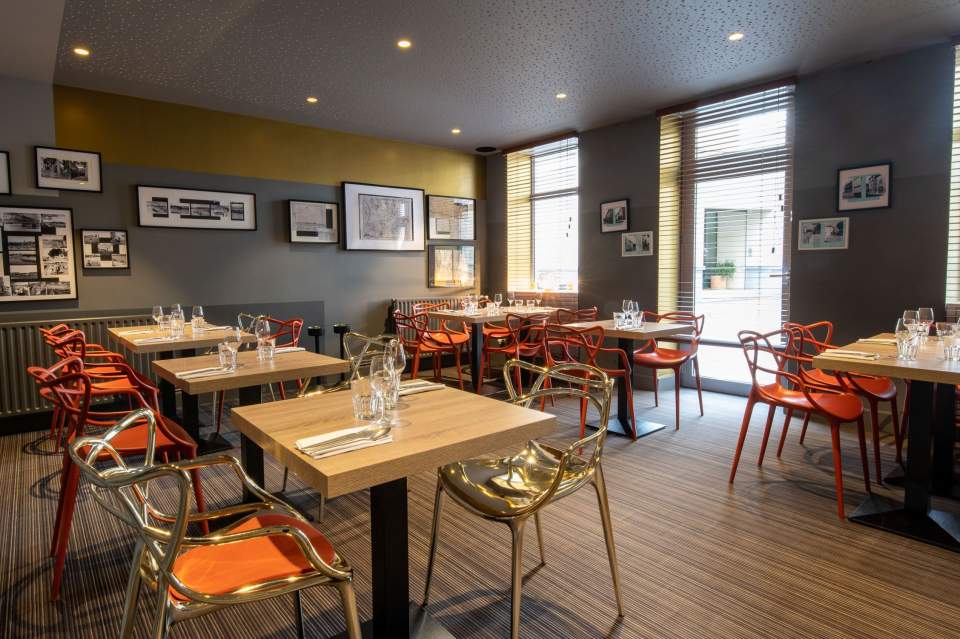 Une salle de restaurant moderne avec des tables en bois, des chaises en métal de couleur argentée et orange, et des murs décorés de nombreuses photos encadrées en noir et blanc.