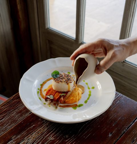 Assiette d'un plat gastronomique composé d'une purée de carotte, d'un poisson nacré et une main verse une sauce dessus
