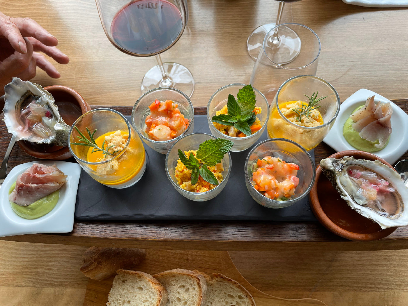 Plateau d'entrées gastronomiques composé de verrines variées, huîtres, tranches de poisson fumé et autres délicatesses, accompagné de pain et d'un verre de vin rouge.