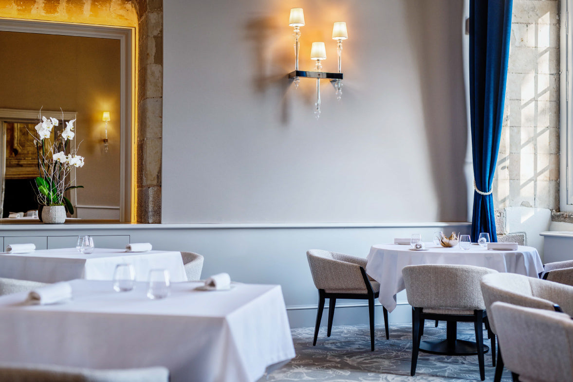 Salle de restaurant élégante avec des tables dressées de nappes blanches, des chaises rembourrées, un éclairage mural raffiné et une décoration florale avec des orchidées blanches, le tout baigné de lumière naturelle.