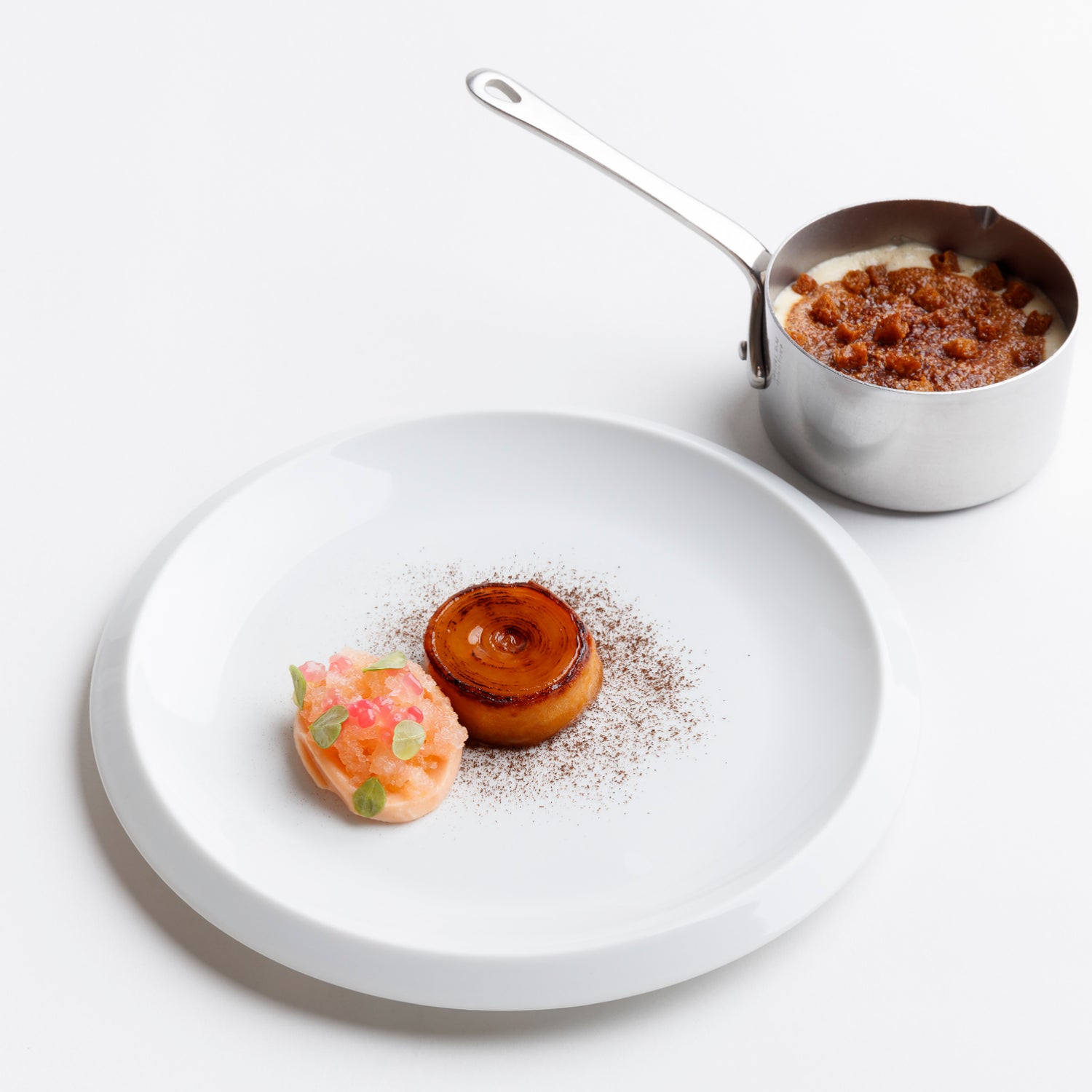 Assiette gastronomique minimaliste avec un dessert composé d'une pomme caramélisée, d'une garniture fruitée et d'une petite casserole contenant un crumble crémeux.