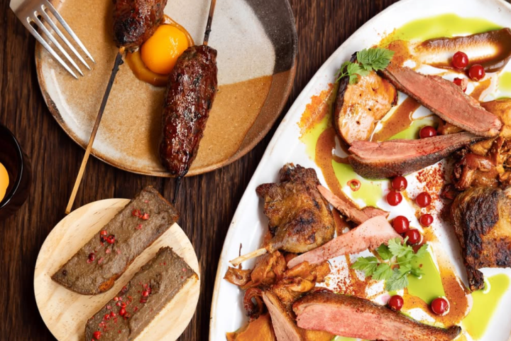 Assortiment de viandes grillées et rôties dressées sur une grande assiette blanche, accompagnées de sauces colorées, d’herbes fraîches et de fruits rouges, avec en complément des brochettes et tranches de pâté servies sur des assiettes en céramique.