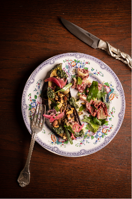Assiette en porcelaine décorative avec un plat composé de légumes grillés, garnis de pickles d'oignon rouge, de noix concassées et d'herbes fraîches, accompagnée d'une fourchette et d'un couteau en métal sculpté.