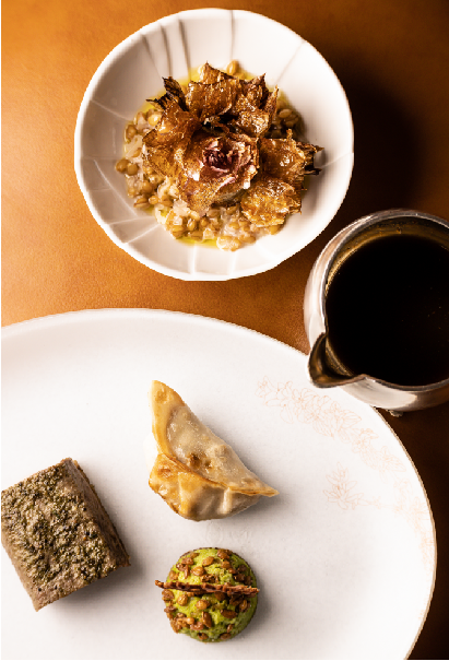 Présentation artistique d'un plat gastronomique sur une assiette blanche, avec divers éléments soigneusement disposés, accompagnée d'un bol contenant une préparation de légumes croustillants sur un lit de grains, le tout accompagné d'une sauce dans une petite cruche à part.