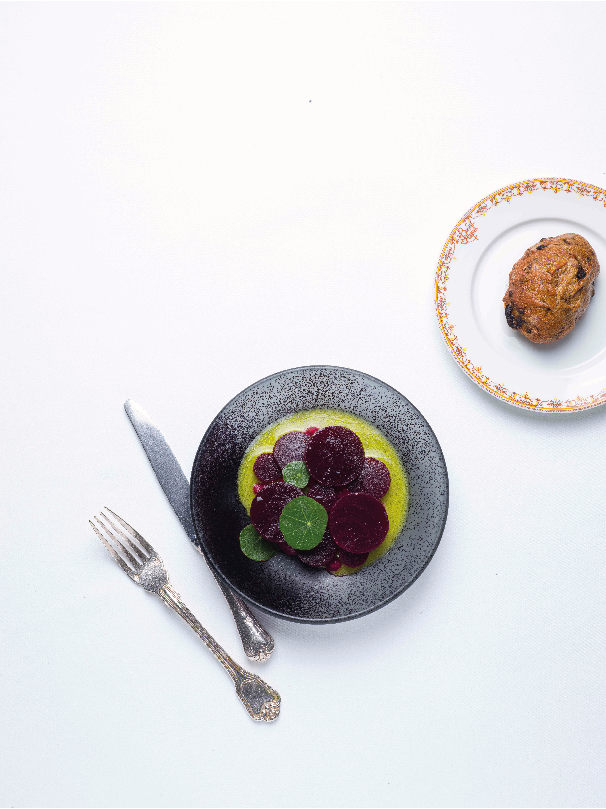 Plat gastronomique minimaliste présenté dans une assiette noire, avec des tranches de légumes colorés disposées sur une purée, accompagné d'un petit pain sur une assiette en porcelaine décorative, le tout sur une nappe blanche immaculée avec des couverts en argent.