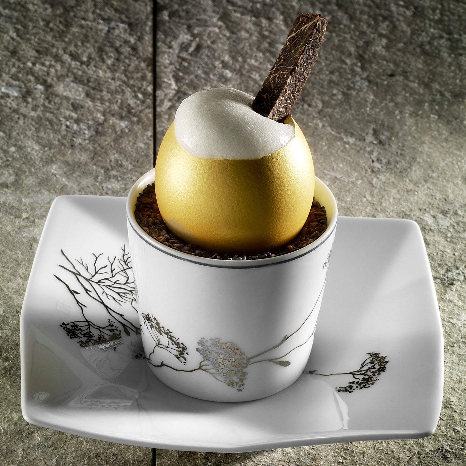 Œuf doré gastronomique garni d'une mousse onctueuse, surmonté d'une lamelle de truffe, présenté dans une tasse décorée posée sur une assiette en porcelaine blanche.
