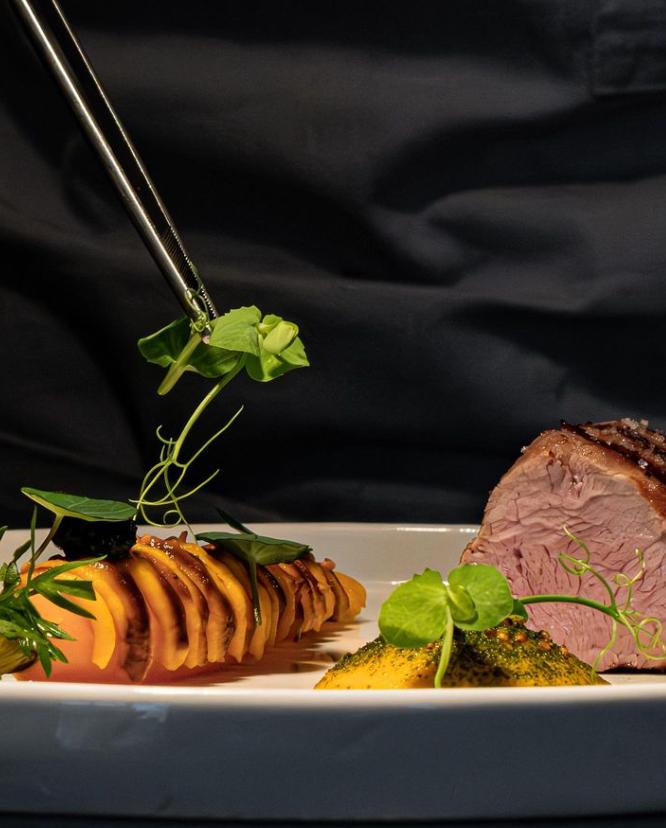 Chef déposant une feuille verte sur un plat de viande élégamment préparé.